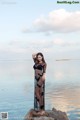 A woman in a black dress standing on a rock by the water.